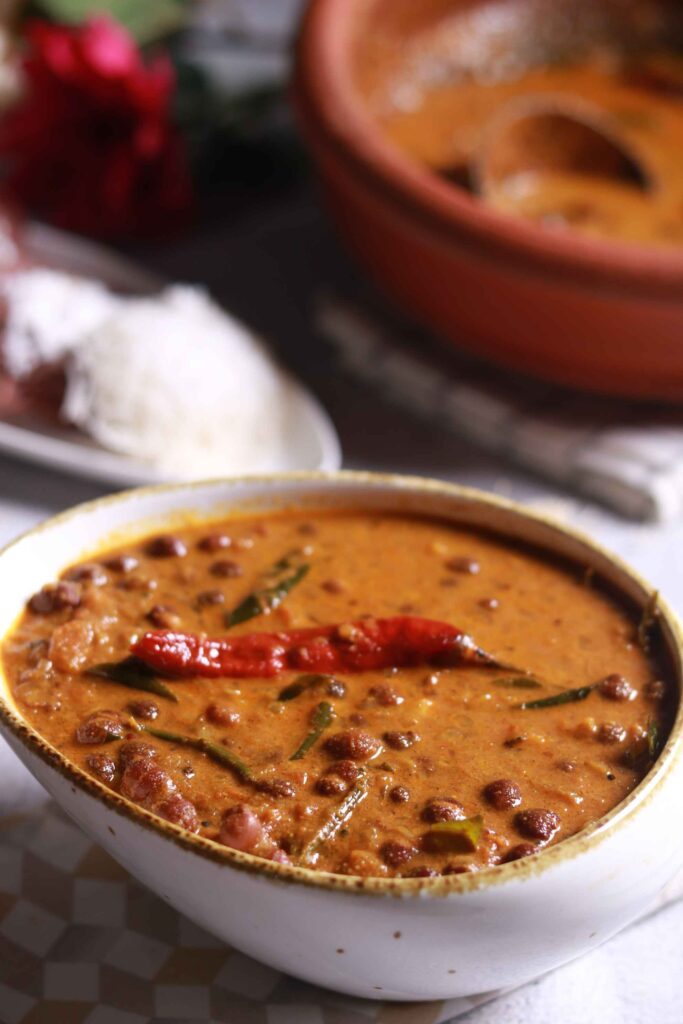 A bowl of Kadala Curry featuring black chickpeas in a rich coconut gravy, garnished with a red chili and curry leaves, served on a table with rice and traditional Kerala dishes in the background.
